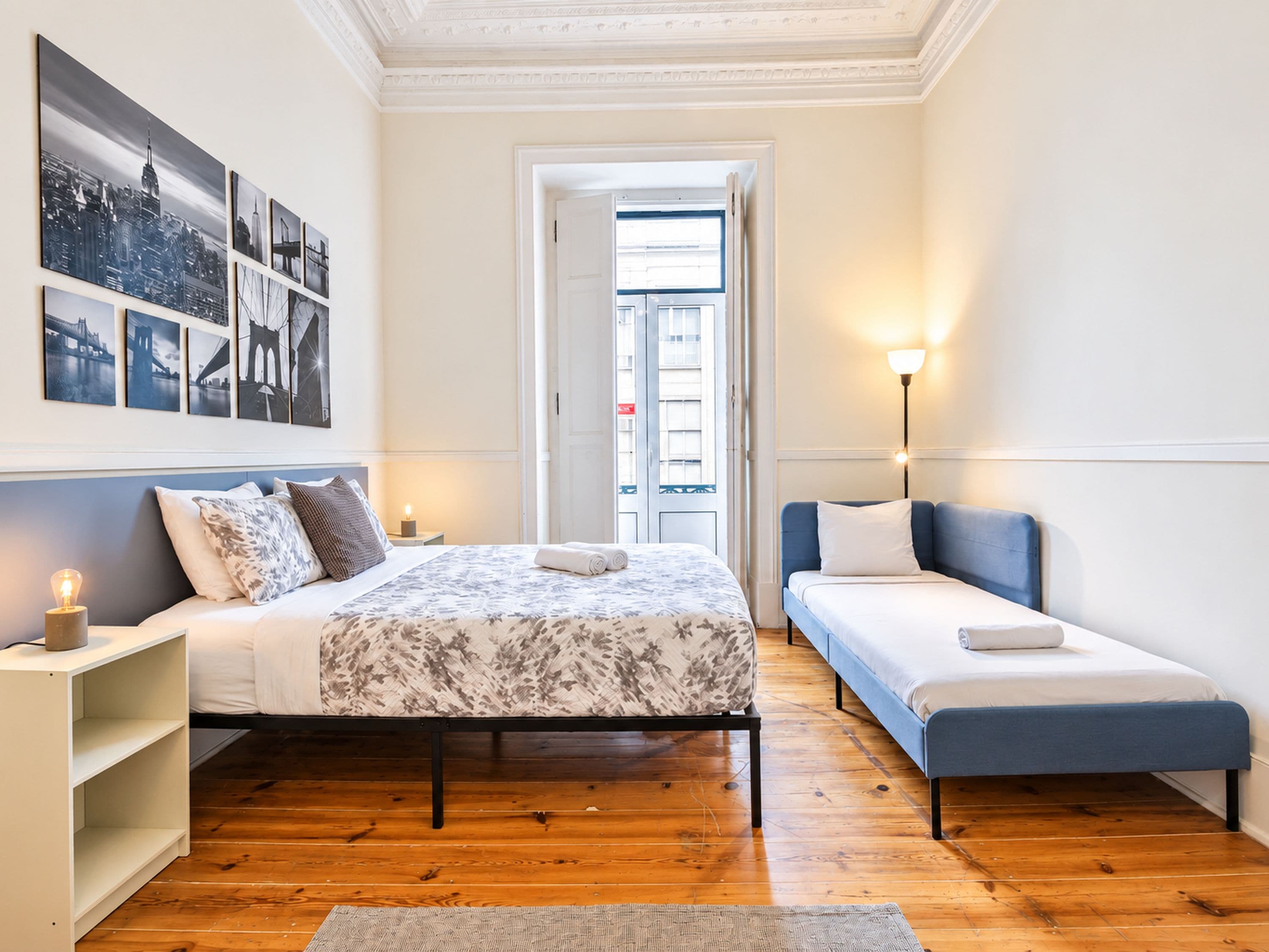 Quarto com cama, mesas de cabeceira, luminária e vista da cidade pela janela.