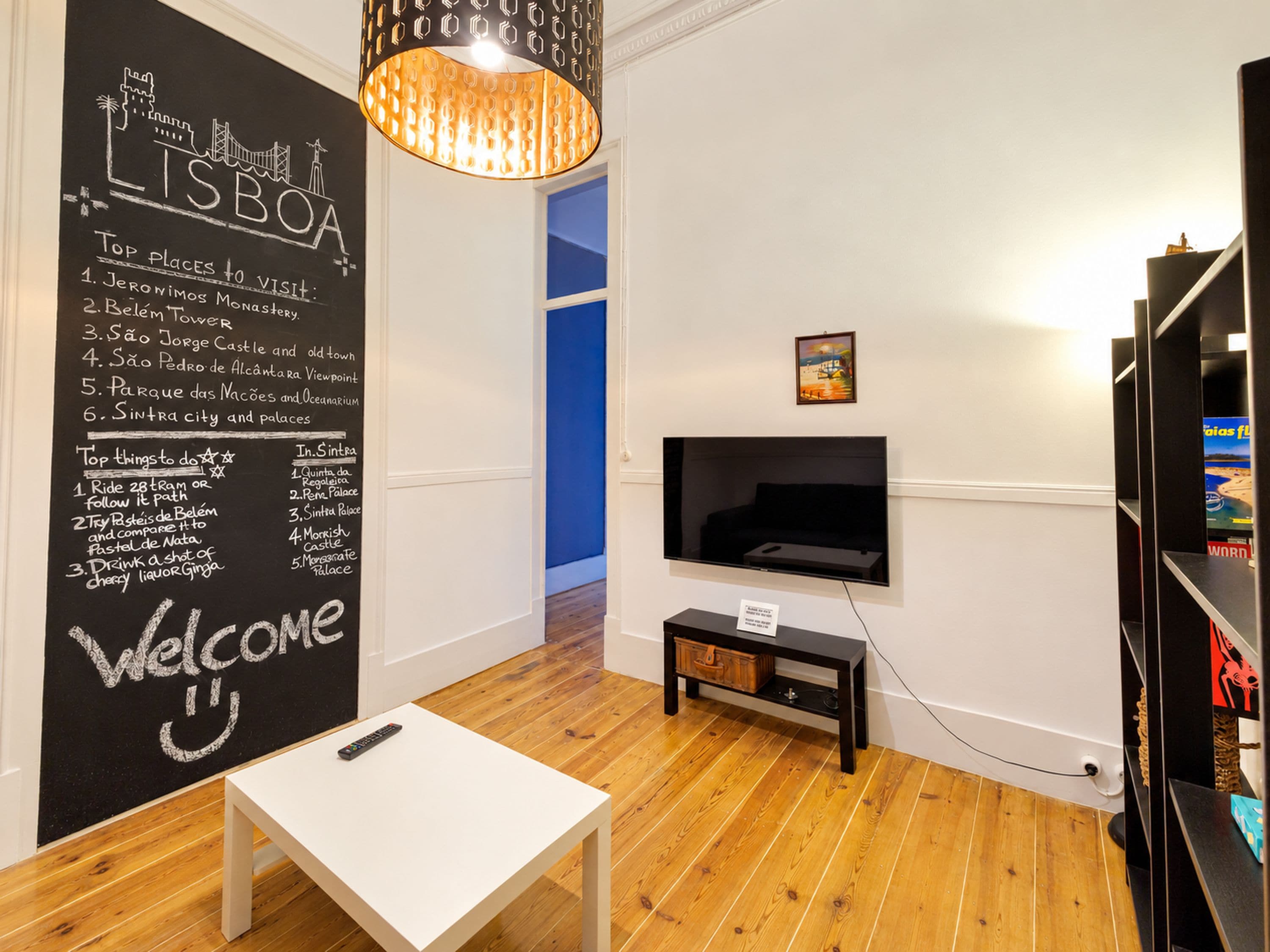 Una habitación con una pizarra que enumera los mejores lugares para visitar en Lisboa, un televisor montado en la pared, suelo de madera y una mesa blanca.