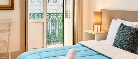 A bedroom with a large bed, a nightstand, a lamp, and a view of buildings through a window.