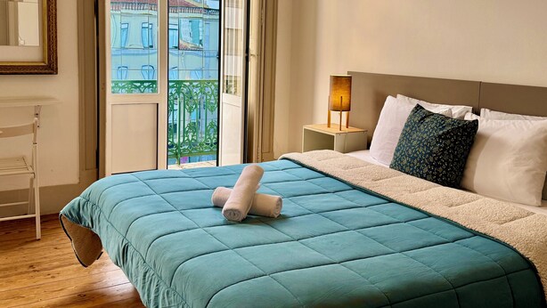 A neatly made bed with a teal duvet and two rolled towels, a bedside table with a lamp, and a view of buildings through a large window. - Ambiente Hostel (Lisbon)