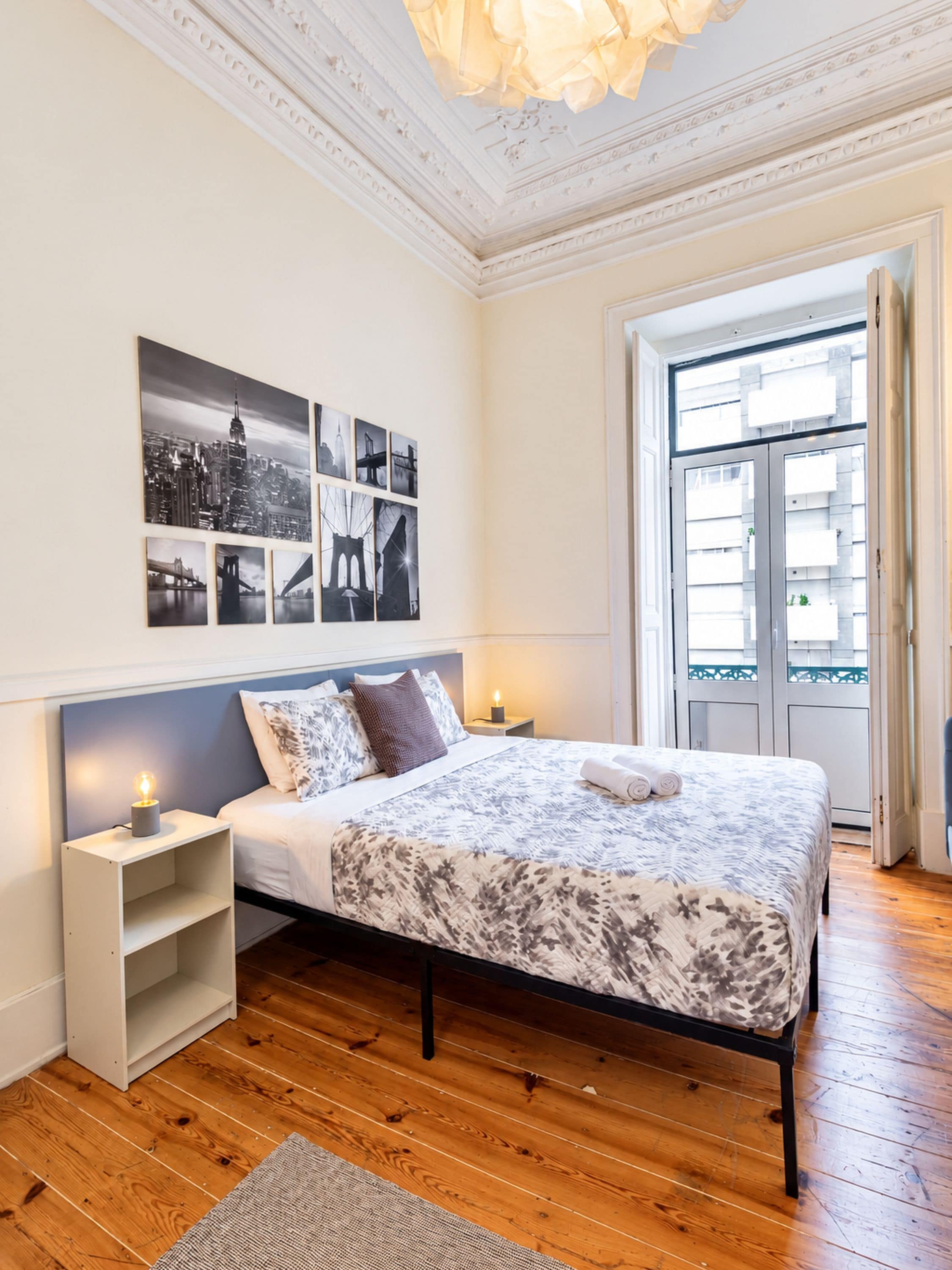 A bedroom with a bed, a nightstand, a wall-mounted picture, and a window with a view of buildings.