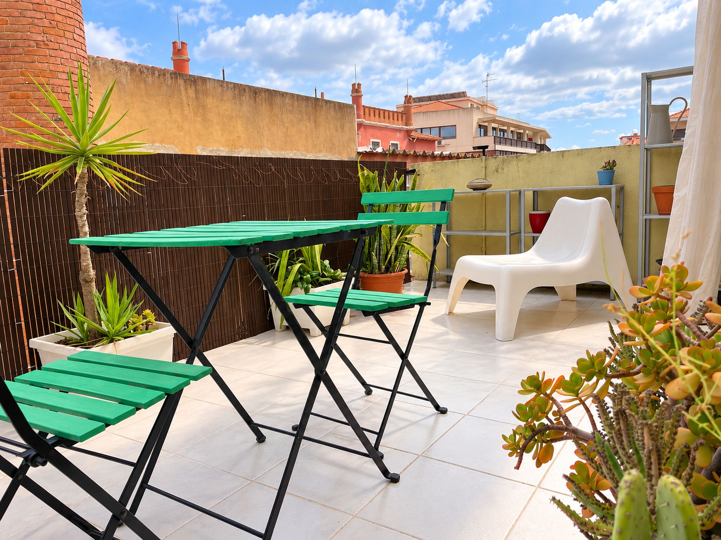 Un patio al aire libre con una mesa y sillas verdes, plantas en macetas y una silla de plástico blanca.