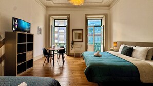 A hotel room with two beds, a desk with chairs, a television, and large windows. - Ambiente Hostel (Lisbon)