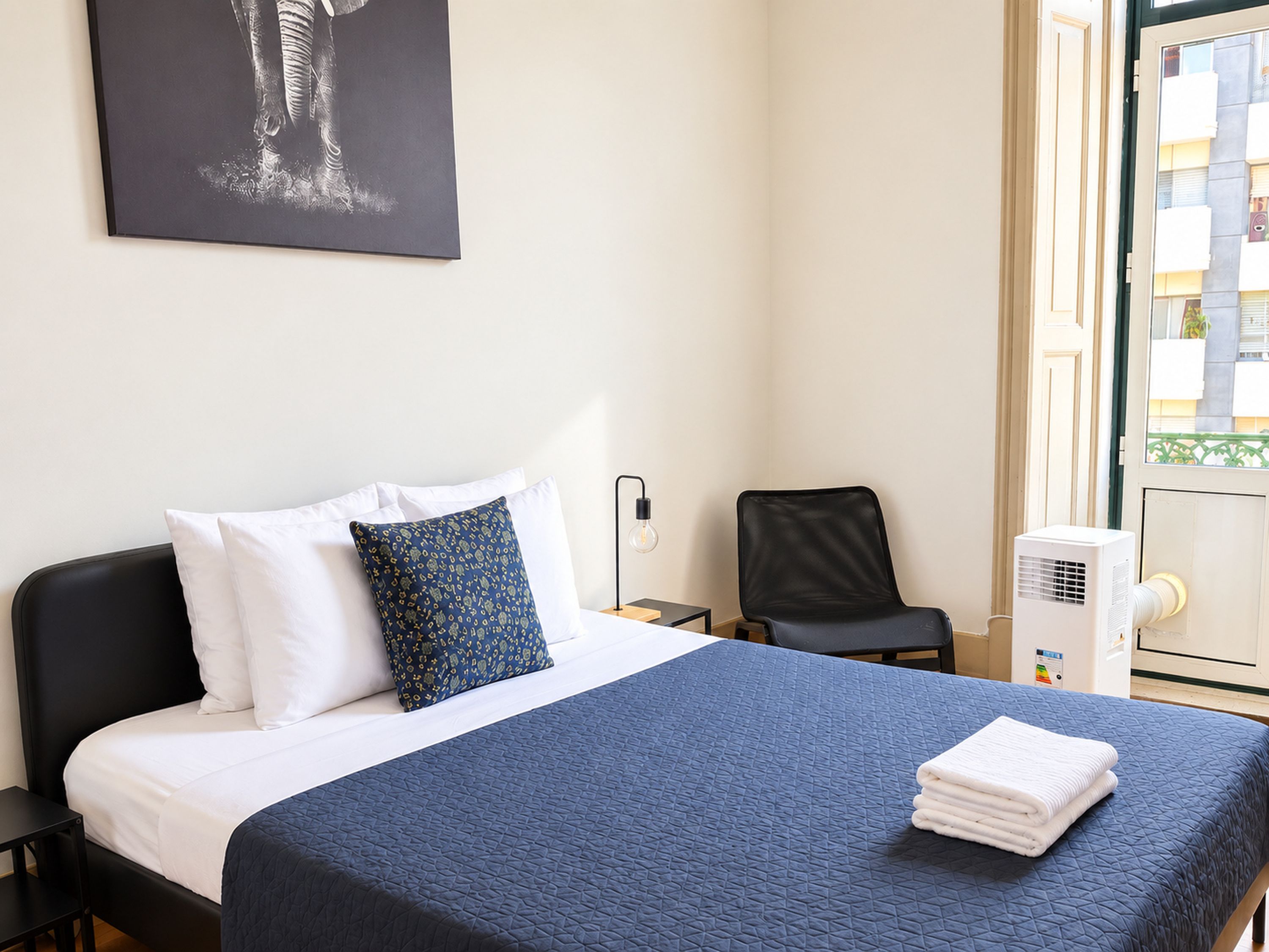 Una habitación de hotel con una cama, una silla, una mesita de noche, una lámpara de pared y un ventanal con vistas a edificios.