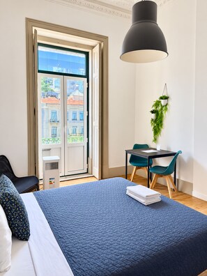 Quarto de hotel com cama, mesa e cadeiras, planta pendurada e vista para edifícios através de uma porta.