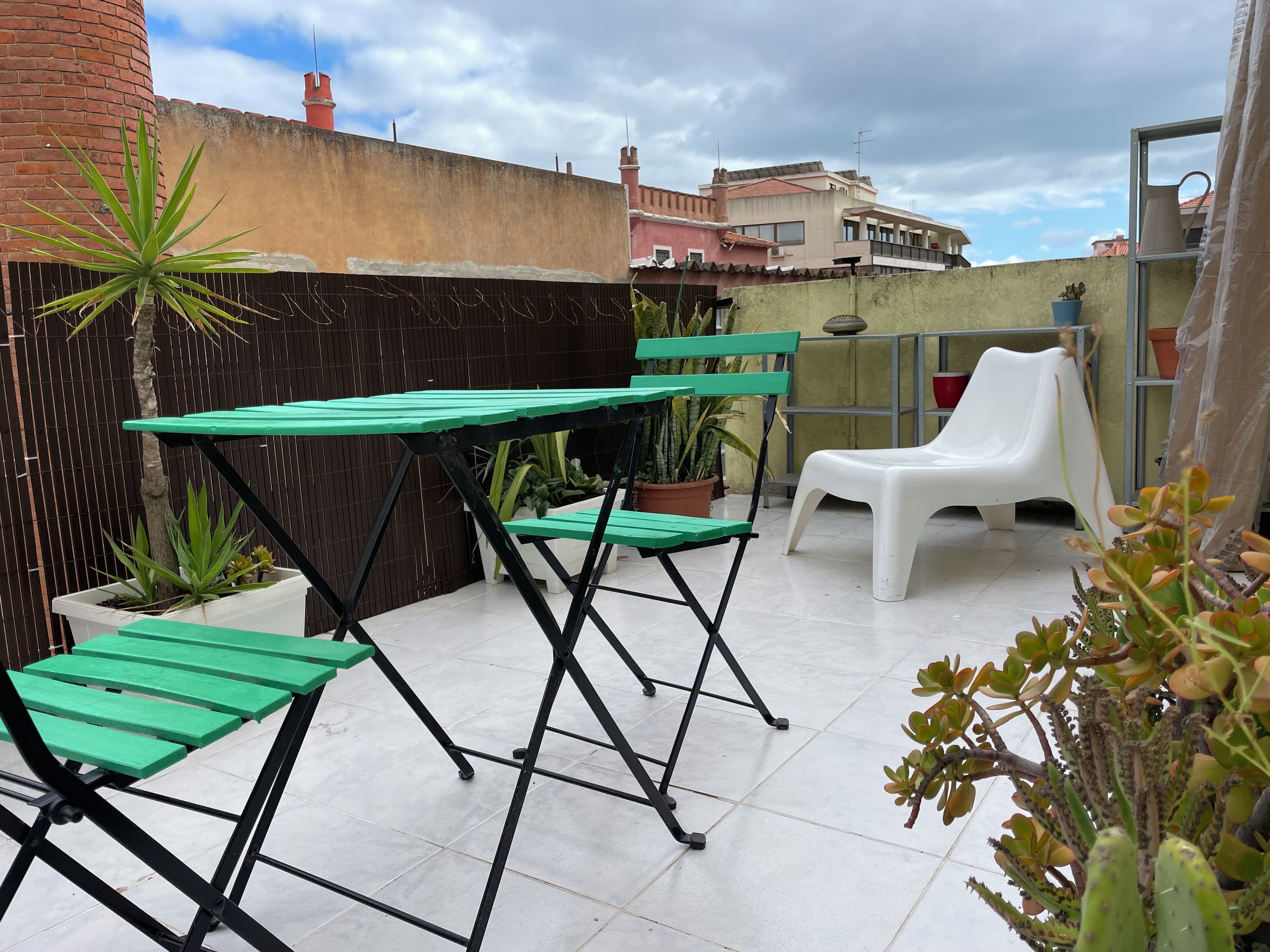 Una terraza en la azotea con una mesa y sillas verdes, plantas en macetas y una silla de plástico blanca.