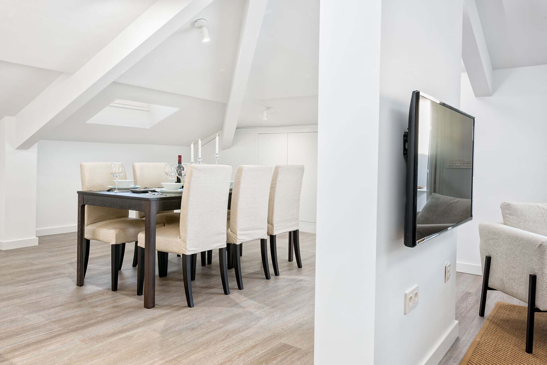A dining area with a dark wooden table, beige upholstered chairs, and a modern TV mounted on the wall.