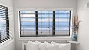 A room with a large window overlooking a beach, a white bed with pillows, and a small shelf with a vase. - Palace 202 (Myrtle Beach)