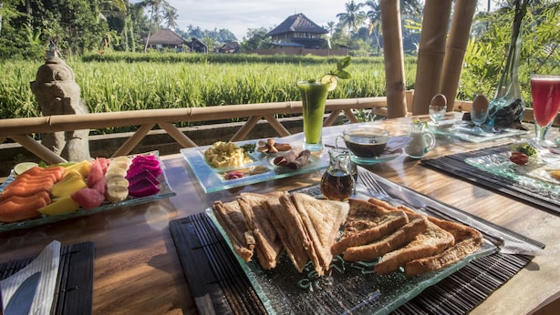 A table set with breakfast items including toast, eggs, and fruit, with a view of a lush garden and a traditional house in the background. - Junjungan Serenity Privite Villa, Spa & Pool (Ubud)