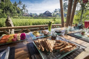 A table set with breakfast items including toast, eggs, and fruit, with a view of a lush garden and a traditional house in the background. - Junjungan Serenity Privite Villa, Spa & Pool (Ubud)