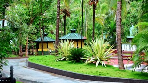Un jardin luxuriant, avec des palmiers et un sentier sinueux menant à un bâtiment surmonté d’un toit végétalisé.