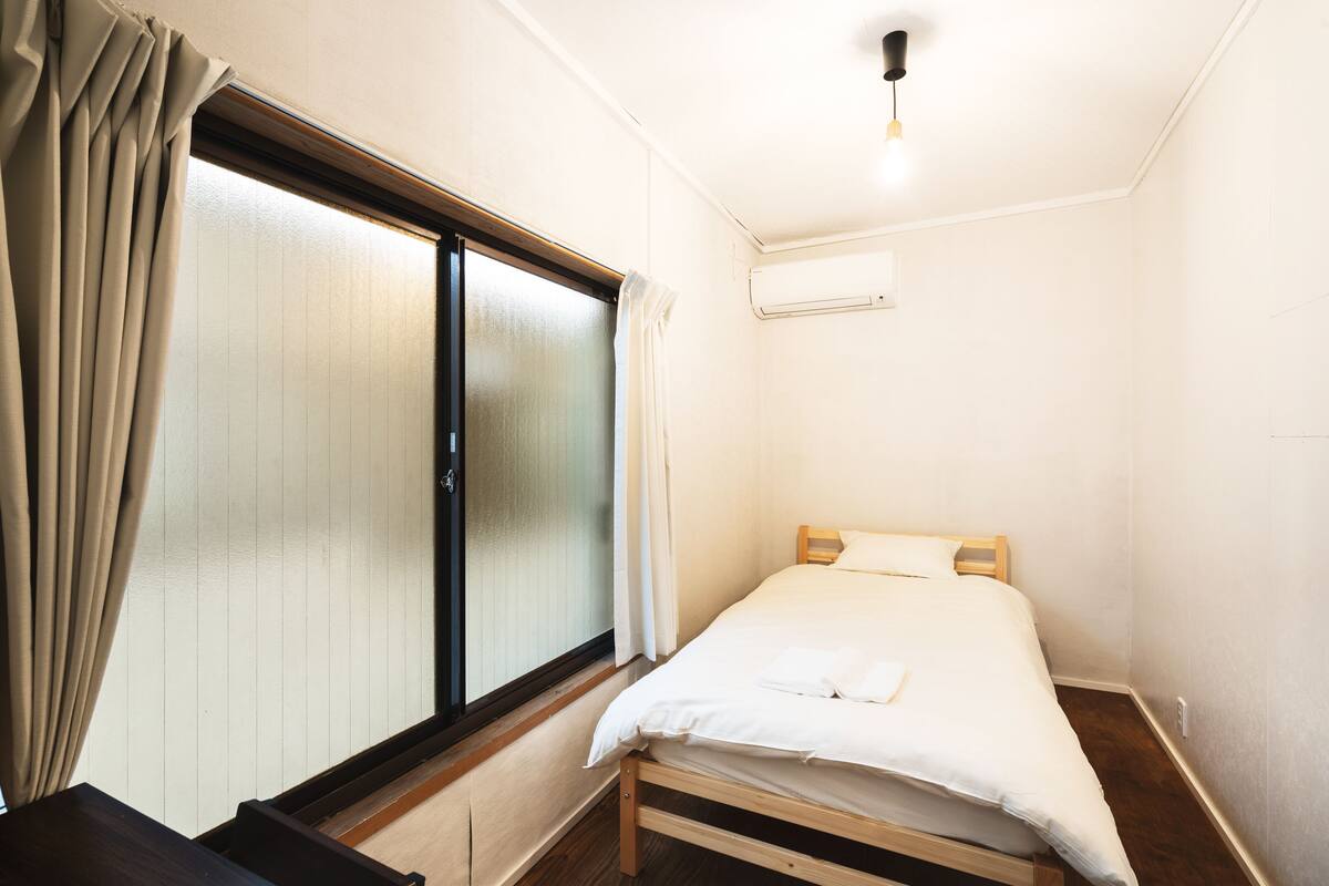 A small, neatly arranged bedroom with a single bed, a sliding glass door, and a ceiling light fixture.