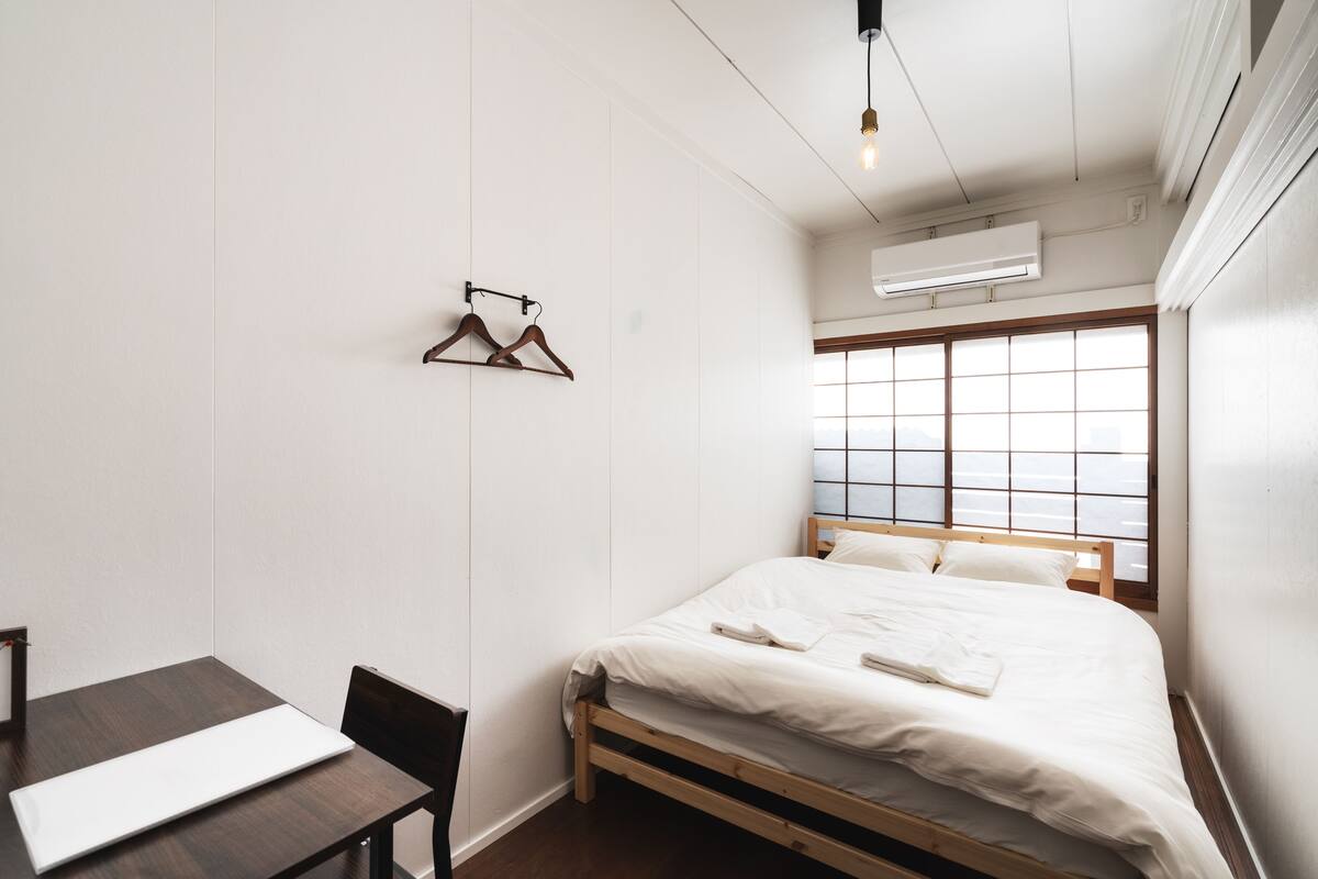 A small, clean room with a bed, a table, and a chair. There are two air conditioning units on the wall, and a window with a grid pattern.