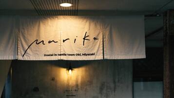 A banner with the name "Marika" and the text "Hostel in castle town Obi, Miyazaki" in a room with a concrete wall and a light fixture.
