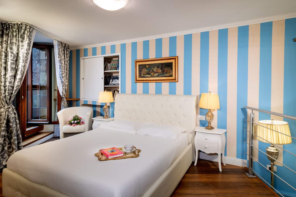 Une chambre d’hôtel avec un grand lit, deux tables de chevet avec des lampes, une chaise agrémentée d’un bouquet de fleurs, une bibliothèque et une fenêtre avec des rideaux.