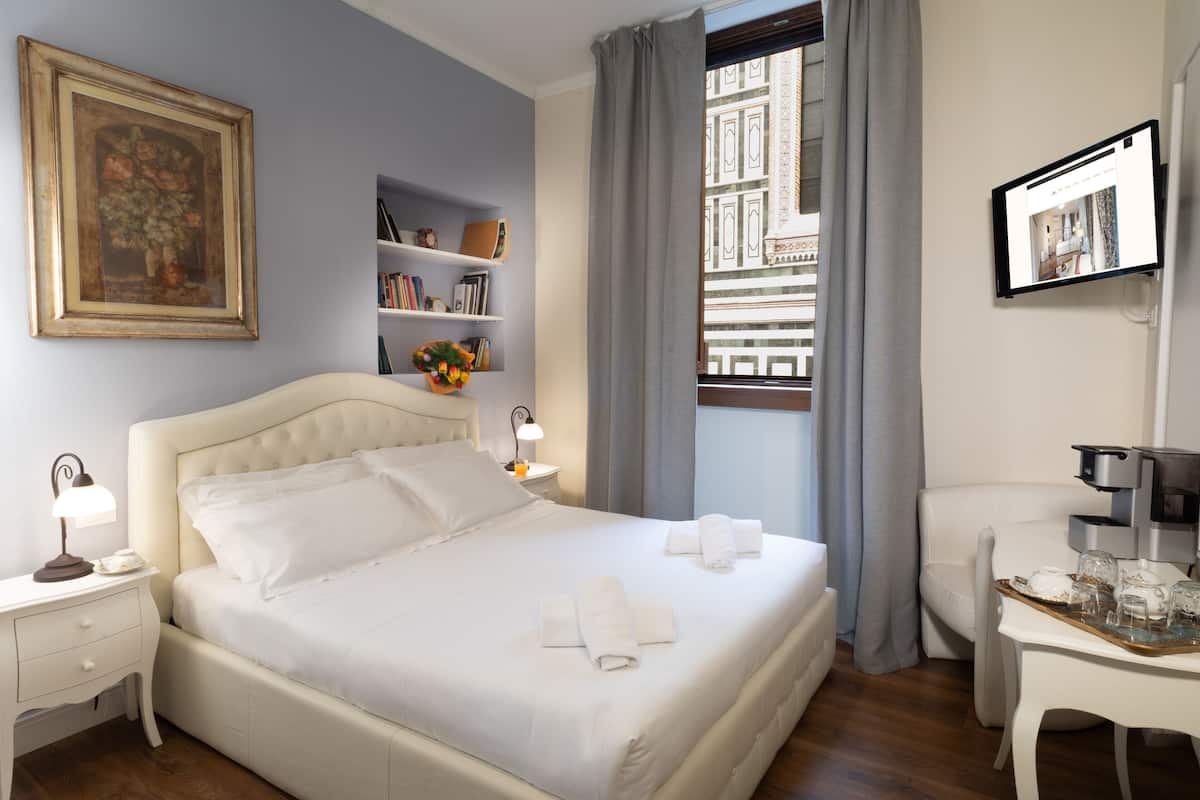 Un lit bien fait, avec du linge de lit blanc, une table de chevet avec une lampe, une bibliothèque et une fenêtre avec des rideaux.