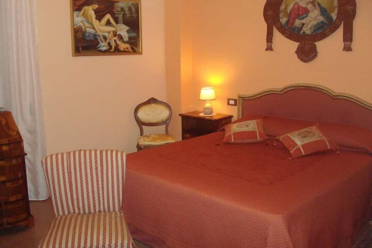 A hotel room with a bed, a chair, a nightstand with a lamp, and two paintings on the wall.