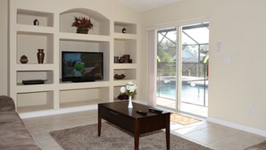Una sala de estar con televisión, estantes, una mesa de centro y un gran ventanal corredizo que da a una piscina.
