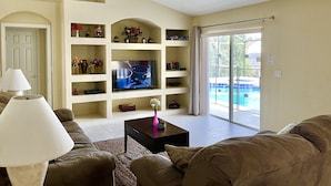 Una sala de estar con televisor, estanterías, ventilador de techo y vista a una piscina a través de un gran ventanal corredizo.