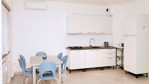A compact kitchen with white cabinets, a gas stove, and a sink. There is a dining table with blue chairs. - Rosa Garden B&B (Mestre)