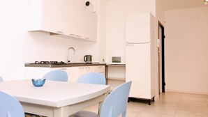 A modern kitchen with white cabinets, a gas stove, a microwave, and a refrigerator. There is a dining table with chairs and a blue bowl on it. - Rosa Garden B&B (Mestre)