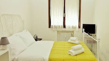 A neatly made bed with a yellow bedspread and white pillows, a bedside lamp, a small table with a lamp, a window with sheer curtains, and a TV on a stand.