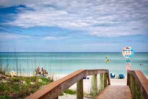 A wooden boardwalk leading to a beach with clear turquoise water, a sign indicating beach access, and a lifeguard warning sign.