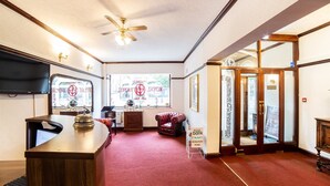 A reception area with a wooden desk, a red carpet, a leather armchair, a ceiling fan, and a view of the outside through a window. - Royal Hotel (Kettering)