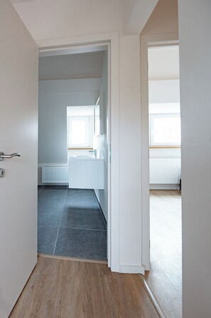 Un couloir menant à une salle de bain dotée d’une fenêtre, d’un radiateur et d’un sol carrelé.