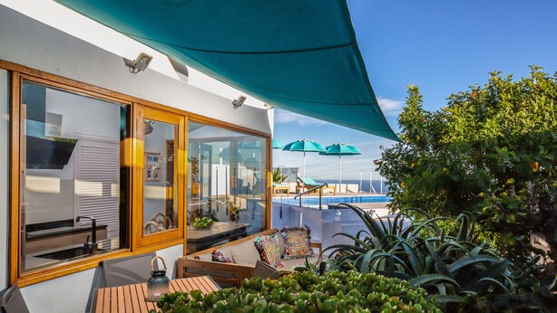 A patio area with a wooden table and chairs, a glass door leading to a room, and a view of a pool with umbrellas.
