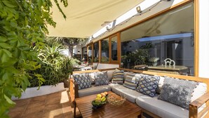 A sunlit outdoor seating area with a wooden bench, cushioned sofa, and a table with fruits.