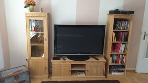 En stue med et fladskærms-tv på et træstel, et glaskab med vinglas, en reol fyldt med bøger, og et lille bord med en vase blomster.