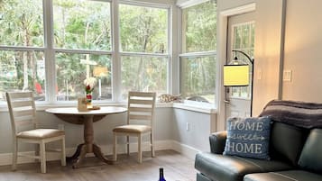 A room with a ceiling fan, a round table with chairs, a sofa with a cushion, and a window with a view of trees.