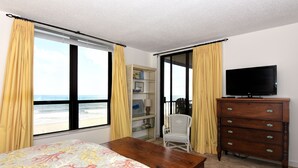 Een slaapkamer met een bed, een dressoir, een televisie en een stoel. Er is een raam met gordijnen, een plank met boeken en een lamp, en uitzicht op het strand.