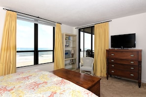 A bedroom with a bed, a dresser, a television, and a chair. There is a window with curtains, a shelf with books and a lamp, and a view of the beach.