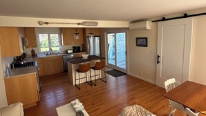 Ein großzügiger Wohnbereich mit Holzboden, einem beigen Sofa, einem Couchtisch und einem Esstisch mit zwei Stühlen. Eine Küche ist sichtbar, mit Holzschränken, einem Kühlschrank und einem Esstisch.