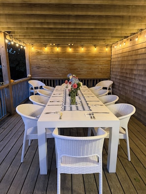 Un área de comedor al aire libre techada con una mesa blanca dispuesta para seis personas, rodeada de sillas de mimbre, bajo luces colgantes.