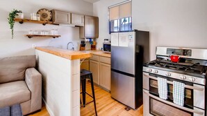 Cocina compacta con refrigerador, estufa y microondas. Incluye un taburete de bar y una pequeña zona de estar con sofá.