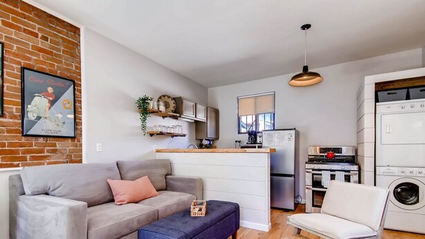 Un espacio habitable pequeño con un sofá gris, un reposapiés azul, una silla blanca, un área de cocina con refrigerador, estufa y lavadora, y una pared de ladrillo con un cartel antiguo.