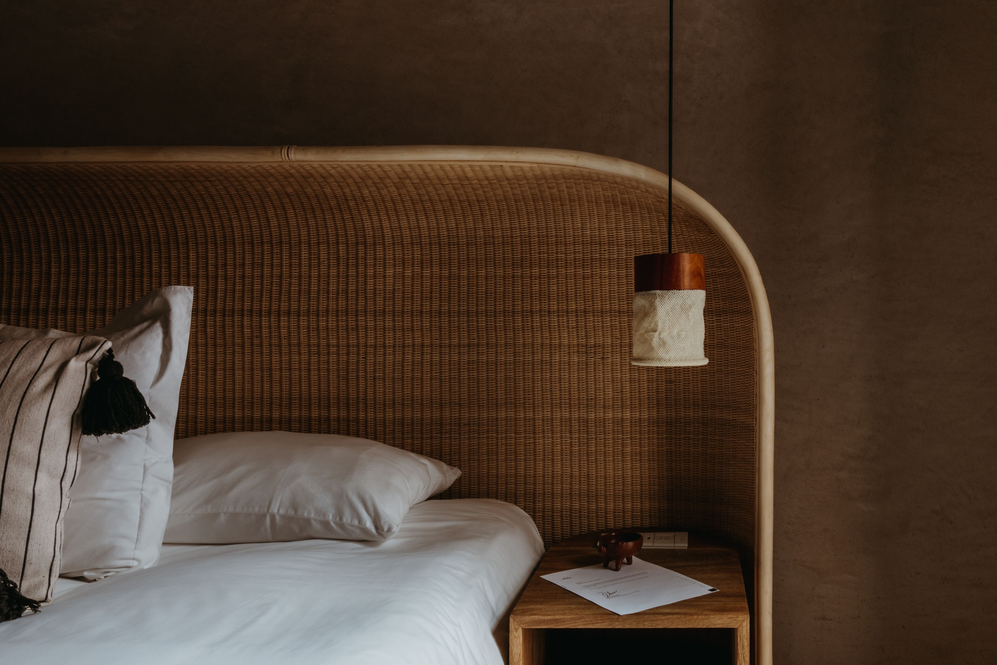 Una cama con sábanas blancas, una mesita de noche de madera, una lámpara colgante y una pared con textura decorativa.