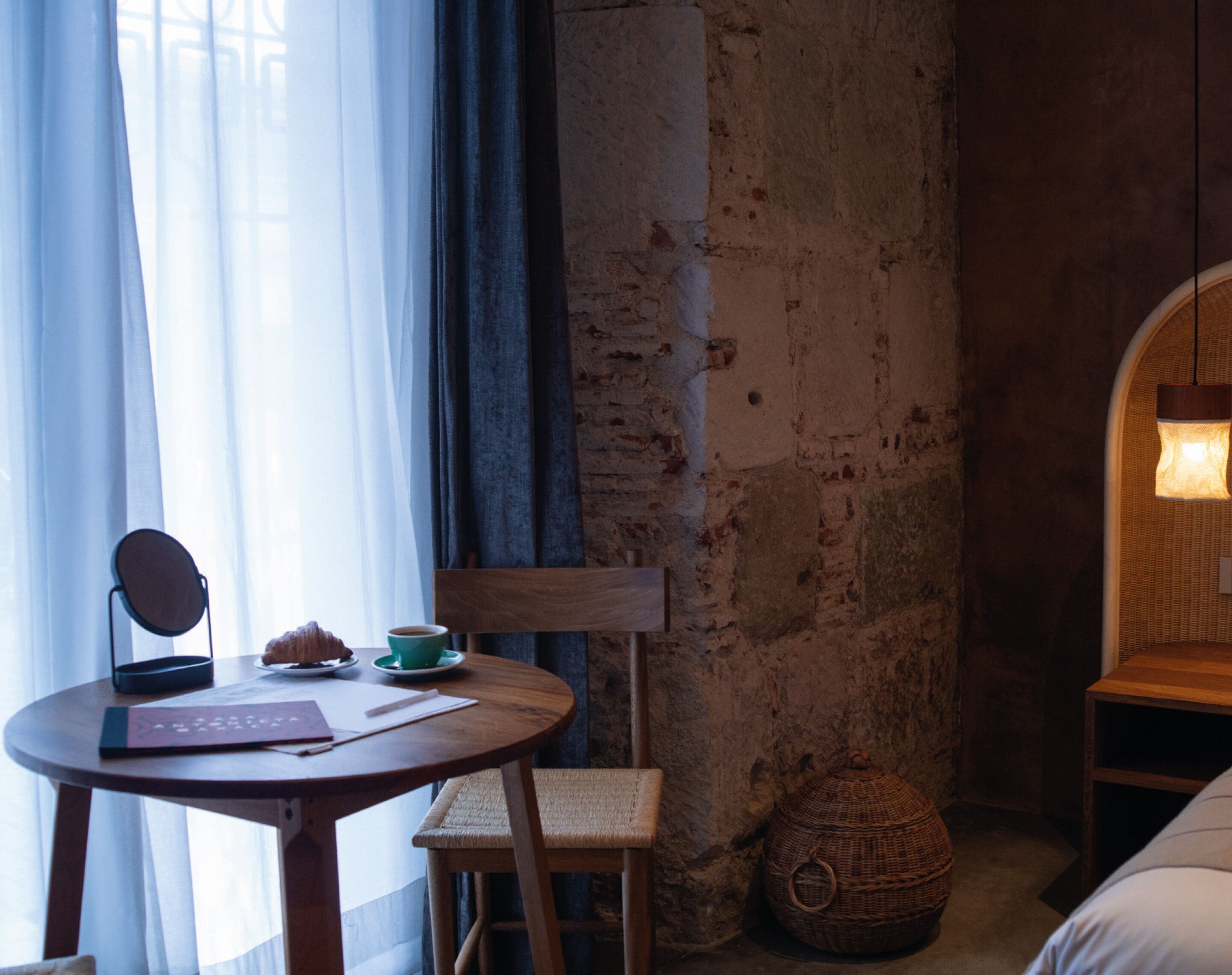 A cozy room with a round wooden table, a chair, a wicker basket, and a bed with a headboard.