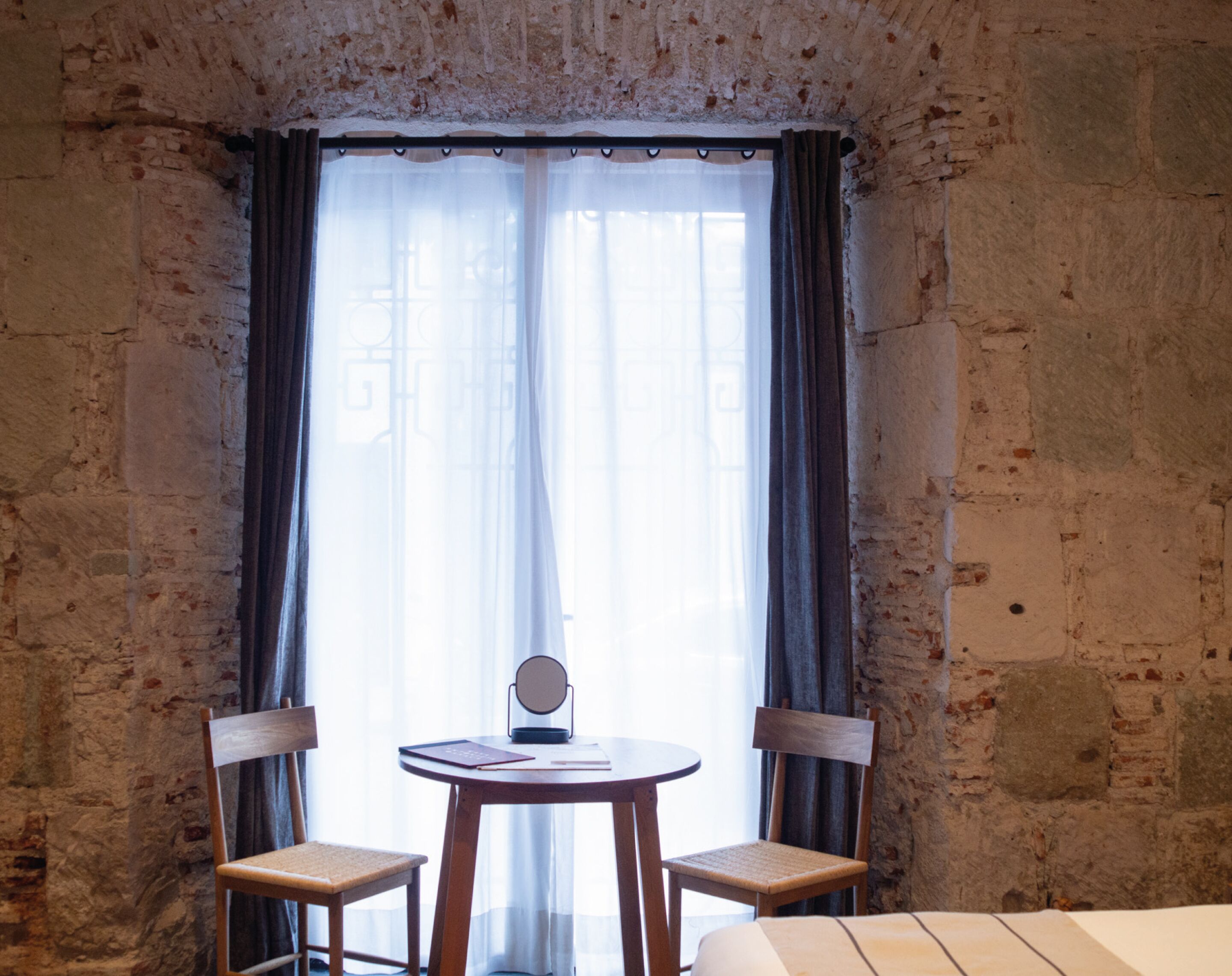 A room with a rustic brick wall, a round wooden table with two chairs, a small round mirror, and a bed with a white headboard.