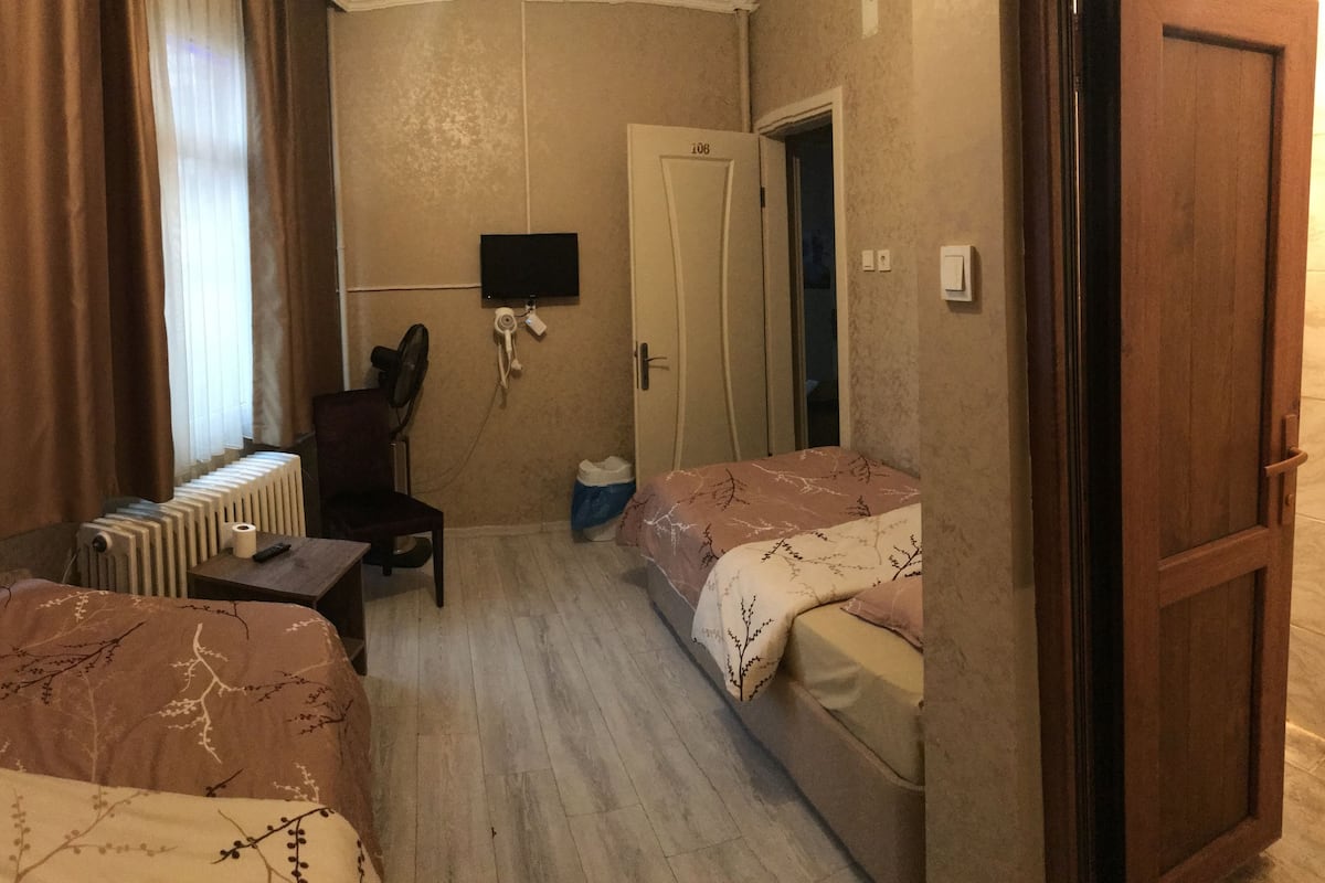 A small hotel room with two single beds, a desk with a chair, a radiator, a television, and a door leading to another room.