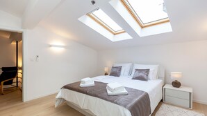 Un dormitorio con tragaluz, una cama con ropa de cama blanca y marrón, una mesita de noche con lámpara y una silla en la esquina.