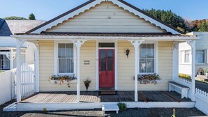 A small, single-story building with a red door, white picket fence, and a porch with flower planters. - Spoil yourself at Strupak Cottage (Nelson City)