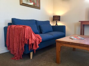 A living room with a blue sofa, a red throw blanket, a wooden coffee table, and a lamp. - Spoil yourself at Strupak Cottage, a quiet retreat close to the city (Nelson City)