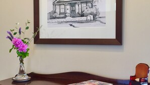 A framed black and white drawing of a house on a wall, a vase with purple flowers on a wooden table, and a book. - Spoil yourself at Strupak Cottage (Nelson City)