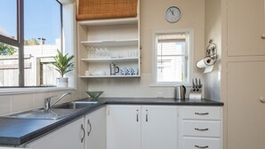 A kitchen with white cabinets, a black countertop, a sink, a clock, and a window with blinds. - Spoil yourself at Strupak Cottage (Nelson City)
