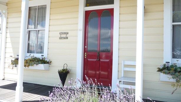 A yellow building with a red door, white window frames, and a porch with white railings. - Spoil yourself at Strupak Cottage (Nelson City)
