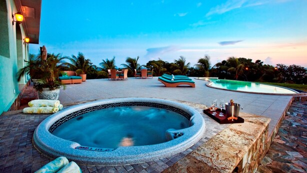 A hot tub surrounded by lounge chairs and a pool, with palm trees and a sunset in the background.